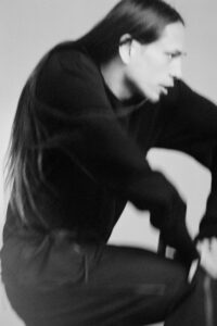A blurred black-and-white scene captures a figure in motion, wearing a dark outfit. Their long hair flows with the movement, and their face is slightly turned, showing focused expression. The composition suggests dynamic motion and energy, creating a sense of fluidity and intensity against a soft, neutral backdrop. The overall atmosphere feels spontaneous and artistic.
