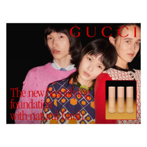 Three individuals pose against a dark background. Their outfits are vibrant and patterned, showcasing a mix of colors and styles. To the side is a display featuring four cylindrical cosmetic products set on a gradient surface, with a warm, inviting glow. The overall scene creates a stylish and contemporary aesthetic with a focus on fashion and beauty.