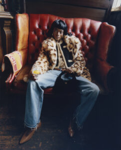 A person with short dark hair reclines on a plush, red-leather couch. They wear a striking leopard-print coat over a patterned shirt and denim jeans, finishing the look with pointed shoes. Holding a small object in one hand, they exude an air of casual confidence and fashion-forward style within a warmly lit, upholstered setting.