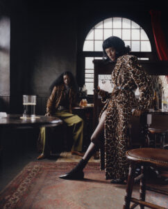 Two individuals are in a dimly lit, stylish room. One stands confidently near a window, wearing a bold, patterned robe and high heels. The other is seated, dressed in a textured top and green pants. The room features wooden furniture, a patterned rug, and a large, arched window that creates a dramatic ambiance.