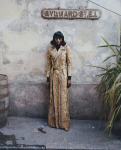 A person stands against an aged, textured wall wearing a long, patterned coat in golden hues. The setting includes a wooden barrel and tall green plants to the side, suggesting an urban environment with rustic elements. The individual gazes directly ahead, exuding a calm, poised demeanor. The overall atmosphere blends vintage charm with contemporary style.