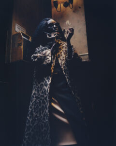 A person stands dramatically under dim lighting, wearing a luxurious leopard print coat. Their posture is expressive, with both hands raised as they look upwards. The setting suggests an urban environment, with dark tones and subtle lighting accents creating a mysterious, theatrical atmosphere. Shadows play across their face, adding a sense of intrigue and sophistication.