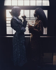 Two women stand facing each other, silhouetted against a large arched window. They wear elegant long coats with intricate patterns. One holds a cup, while the other gestures with her hand. Light streams through the window, casting soft shadows. Their conversation appears intimate and relaxed, creating a serene, contemplative atmosphere enriched by the play of shadow and light.