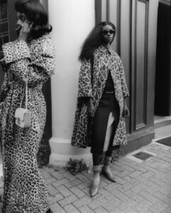 Two individuals stand confidently on a sidewalk, exuding style and attitude. Both wear leopard-print coats; one person is speaking on the phone with a small purse, while the other sports sunglasses and a layered skirt. The setting features elegant architectural elements, including a prominent column and wooden panels, adding a classic urban vibe to the scene.