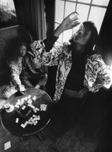 A monochrome scene captures two individuals in a cozy setting. One person lounges on a bench, wearing a printed coat, while the other stands, drinking from a glass. A table with playing cards, a cup, and various items adds interest. Natural light filters in through a window, creating a moody atmosphere with sharp contrasts and textures.