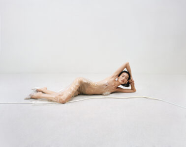 A person is lying on a light, expansive floor, surrounded by minimal surroundings. The individual is partially wrapped in translucent material, creating an ethereal and serene atmosphere. Their eyes are closed and arms positioned thoughtfully, enhancing the sense of calm and introspection. The setting is stark, with muted colors that emphasize the tranquility and simplicity of the scene.