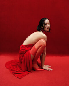 A figure with shoulder-length dark hair is posed on the ground draped in a long, flowing red fabric. They appear to be wearing red stockings and are barefoot. The background and the surface they sit on are both a deep red, creating a monochromatic setting. The overall composition emphasizes the striking contrast and elegance of the figure.