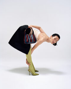 A person bends over, holding a multicolored striped bag. They wear a light beige tank top, black skirt, and lime green tights, paired with pointed high heels featuring a gold chain detail. Their hair is slicked back, and they sport long earrings. The overall aesthetic is modern and fashion-forward, emphasizing bold colors and unique styling.