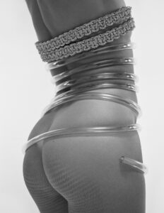 A monochrome composition features a close-up of a person from the side, adorned with intricately textured fabric around their waist. Transparent tubes elegantly coil around them, running from the torso to the thighs, creating a dynamic interplay between the smooth materials and the subtle, patterned stockings. The minimalistic setting enhances the focus on form and texture.