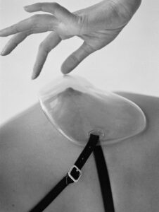 A delicate hand gracefully extends downward, hovering above a smooth, elliptical, clear object attached to a person's back with a thin, black strap and silver buckle. The composition is minimalistic, emphasizing the contrast between the soft hand and the structured object. Shadows and light interact subtly, creating an intriguing interplay of textures and forms.