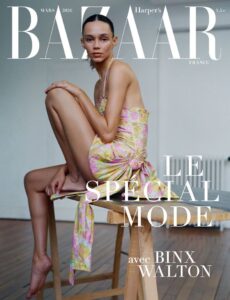 A woman sits sideways on a simple wooden stool in a minimal interior, her body softly curved and poised. She wears a delicate floral dress with thin straps and a tied detail falling down. Her expression is calm and direct. Bare feet rest lightly, natural light highlighting smooth skin, muted tones, and an intimate, understated elegance in a quiet room.
