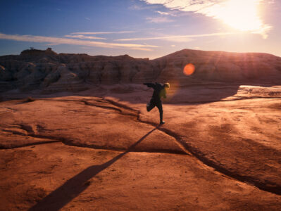 A solitary figure sprints across a vast, arid landscape bathed in warm, golden light. The rugged terrain, marked by cracks and rocky formations, stretches out under a vivid sky. Sunlight streams through wispy clouds, casting long shadows and creating a dramatic play of light and shadow. The scene evokes a sense of freedom and boundless energy.
