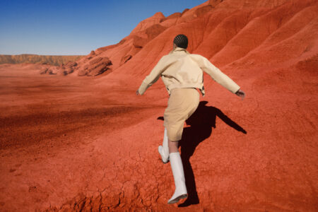 A person dressed in a light beige outfit and white boots walks across a vast, rocky landscape of vibrant red hues, under a clear blue sky. The terrain is textured with cracks and small formations, creating striking contrast. The scene evokes a sense of exploration and adventure, highlighting the dramatic natural beauty and stark colors of the environment.