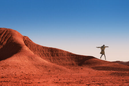 A person is captured mid-jump on a striking red desert landscape, characterized by smooth, rolling hills and vibrant terrain. The backdrop features a clear, gradient sky transitioning from pale blue at the horizon to a deeper blue overhead. The scene evokes a sense of freedom and exploration amidst the stark, expansive beauty of the arid environment.