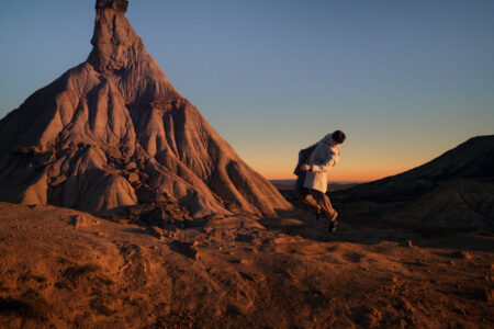 A person, wearing a light-colored jacket, is captured mid-movement against a rugged desert landscape. The backdrop features a distinct, pointed rock formation with textured surfaces, illuminated by warm sunset hues. The sky transitions from deep blue to orange near the horizon, creating a dramatic and serene contrast. A sense of adventure and vastness pervades the scene.
