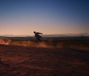 Amid a desert landscape, a person is moving dynamically, slightly leaning forward as they walk through a reddish sandy terrain. The sky transitions from a warm golden hue near the horizon to a deep blue above. Dust swirls around their feet, highlighting the arid and expansive environment under a setting or rising sun.