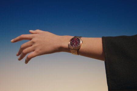 A hand is held against a smooth gradient background shifting from deep blue to soft beige. On the wrist, a sleek watch with a metallic band and a circular face marked by large numbers adds sophistication. The design contrasts the hand's natural tones with the timepiece's metallic shine, creating an elegant and modern aesthetic.