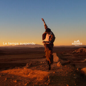 A person dressed in formal clothing appears carefree and dynamic against a vast desert landscape. The sun casts a warm, golden glow over the scene, illuminating the surroundings with soft light. The horizon stretches far, with distant mountains visible under the clear sky. The moment captured conveys movement, exploration, and a sense of freedom amid nature.