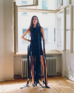 A person stands confidently in a sunlit room, wearing a distinctive black outfit with long, flowing strips cascading towards the floor. The setting features an open window, with natural light illuminating the wooden floor and white radiator. The scene conveys a sense of bold fashion within a minimalist, airy environment, merging modern style with an urban backdrop.