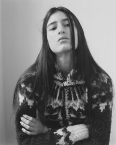 A person gazes forward with a neutral expression, their long hair falling straight over their shoulders. They wear a patterned, knitted sweater with distinct geometric designs. The lighting is soft, and the background is plain, creating a serene, minimalist atmosphere. Arms are crossed, adding a touch of casual defiance or contemplation to the overall composition.
