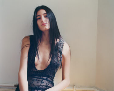 A person with long, dark hair is seated against a cream-colored wall. They wear a sheer, black dress adorned with intricate lace and beadwork. The neckline is deep, adding elegance to the look. The individual gazes forward with a serene, contemplative expression, while soft lighting enhances the peaceful and introspective mood of the setting.