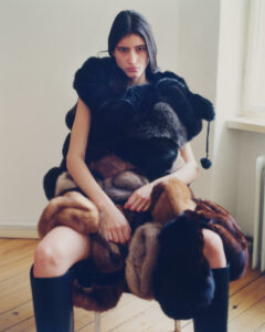 A person with long dark hair sits on a chair in a minimalist room. They are draped in a voluminous, multi-textured fur coat with rich, dark hues. Sunlight streams in from a nearby window, casting soft shadows on the wooden floor. Their expression appears contemplative, and they wear tall, sleek black boots, adding to the striking ensemble.