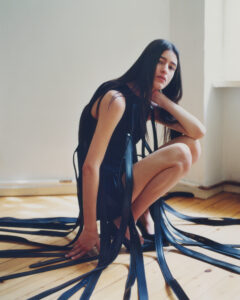 A person with long dark hair sits on a wooden floor in a softly lit room. They are wearing a black outfit with multiple long, ribbon-like extensions that spread around them. The person rests one elbow on their knee, with a pensive expression. Light streams in from a window, casting gentle shadows on the floor.