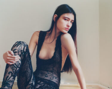 A contemplative individual with long, straight hair gazes outward while seated in a corner. They wear an intricately designed, sheer black outfit adorned with ornate patterns and beading. Sunlight softly filters into the room, casting subtle shadows on the plain walls. The atmosphere conveys a sense of quiet introspection and elegance.