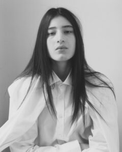 A monochrome portrait features a person with long, straight hair cascading over a crisp white button-up shirt. They maintain a neutral expression, with a soft, natural look. The lighting is soft, casting gentle shadows that accentuate the serene and contemplative mood. The background is plain, directing focus entirely on the central figure's expression and attire.
