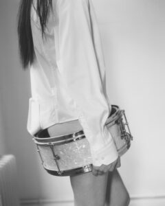 A monochrome scene features a person with long hair, wearing a long-sleeved shirt, and holding a drum around their waist. The backdrop appears minimalist, with an even-toned wall and soft lighting casting gentle shadows. The overall composition blends the crisp texture of the shirt with the smooth surface of the drum, creating a striking artistic contrast.