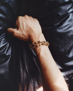 A hand adorned with a gold chain bracelet rests against a backdrop of textured black material. The lighting casts subtle shadows and highlights, emphasizing the contours of the wrist and the intricate details of the jewelry. The black surface behind adds depth and contrast, creating a sophisticated and elegant visual composition that draws attention to the golden accessory.