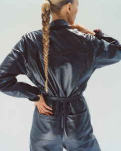A person with long blonde hair in a braid stands with their back facing, dressed in a shiny, dark, leather-like outfit. The outfit features a high collar and pockets, with one hand placed on the hip and the other adjusting the collar. The light reflects off the fabric, highlighting its texture and elegant design.