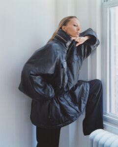 A person stands pensively by a window, wearing an oversized black leather jacket with a high collar. Their left arm rests on a knee propped on a white radiator, while the right hand lightly touches their chin. The setting is a bright room with white walls, allowing natural light to cast soft shadows, highlighting the jacket’s texture.