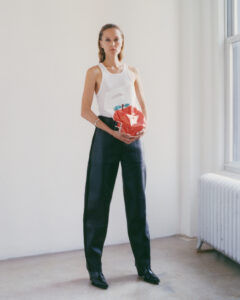A person stands confidently in a minimalist, bright room. They're dressed in a white sleeveless top and loose black trousers, holding a shiny, red, apple-shaped handbag. The sleek black shoes and belt add sophistication. Natural light streams through a large window, enhancing the clean, modern aesthetic of the scene. A white radiator is visible nearby, completing the setting.