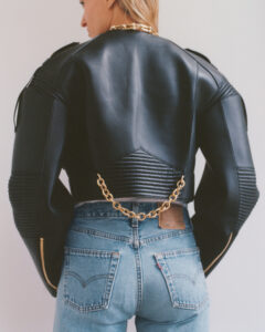 A person with short blonde hair is seen from behind, wearing a black leather jacket featuring quilted detailing and gold chain accents. The jacket has structured shoulders and zipper details. Paired with light blue denim jeans, the ensemble highlights both rugged and sophisticated elements. The overall composition emphasizes a fusion of modern fashion trends and classic styles.