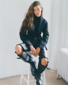 A person with long, wavy hair sits confidently on a white stool in a bright room. They wear a sleek black blazer over a dark turtleneck sweater, paired with black leather pants and knee-high boots. The overall pose and attire suggest a sense of modern, poised fashion. The minimalistic background highlights the individual's stylish presence.