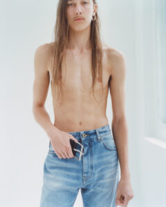 A person with long, straight hair is standing in a well-lit space. They are shirtless, wearing high-waisted, faded blue jeans. An accessory is held in one hand at the waist. Their expression is neutral, featuring a mustache and one visible earring. The minimalist background highlights the relaxed and straightforward style, focusing on the casual attire.