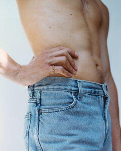 A shirtless person is seen from the waist up, wearing light blue jeans. Their hand rests on their hip, adorned with a gold ring featuring a unique design. The lighting emphasizes the contours of their torso and the texture of the denim, creating a contrast between the skin and the fabric, set against a simple background.