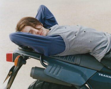 A person with short, tousled hair reclines comfortably on a motorcycle seat. They wear a layered outfit consisting of a gray, hole-punched short-sleeved top over a blue long-sleeved shirt. Their arms rest behind their head, conveying a relaxed demeanor. The setting is an open, sandy area, with soft, natural lighting creating a tranquil atmosphere.