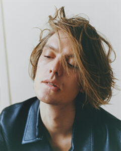 A person with tousled, light brown hair appears relaxed and contemplative, with eyes partially closed. The head is tilted slightly back, capturing a serene expression. Wearing a dark leather jacket with visible stitching along the collar, the individual exudes a casual yet stylish vibe. The backdrop is softly blurred, adding a gentle, introspective atmosphere to the scene.