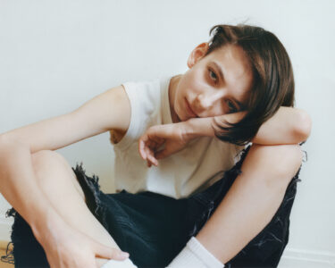 A contemplative youth is seated casually with head resting on a bent arm. The person wears a sleeveless white top and distressed black jeans, paired with white socks. Hair falls across the forehead. The posture and expression convey a sense of introspection and calm, set against a minimalist background of soft whites and neutrals.