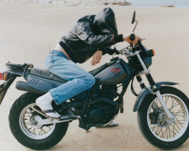 A person wearing a hooded leather jacket and jeans leans forward on a parked motorcycle on a sandy surface. The rider is sitting casually on the bike, with their head lowered and hand gripping the handlebar. They wear white socks with sandals, and the background shows a blurred, barren landscape with a few rocks visible.