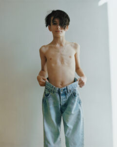 A young person with dark, tousled hair stands confidently, wearing oversized light blue jeans held at the waist. Their upper body is bare, and a delicate chain necklace adds a touch of elegance. A neutral, softly lit background creates a minimalist effect, highlighting the contrast between the youth's slender frame and the too-large denim.