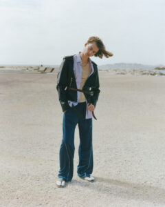 A person stands on a sandy, open landscape with their head tilted, and hair blown by the wind. They wear a layered outfit consisting of a jacket, unbuttoned shirt, and loose blue trousers. The background includes distant blurred structures and a calm sky. The overall vibe is relaxed and contemplative, suggesting a casual fashion photoshoot.