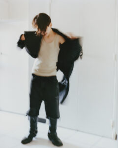 A person stands in a softly lit space, moving slightly, causing motion blur. They are wearing a light-colored tank top, dark shorts, and knee-high boots. A dark jacket is draped over their shoulders. Their hair is tousled, partially obscuring their face. The setting appears minimalist with smooth, white surfaces around them, suggesting a modern environment.