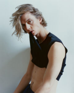 A person with light hair, styled messily, gazes ahead with an intriguing expression, biting a black, sleeveless top. The pose is slightly tilted, revealing a toned arm and torso with visible freckles. The background is a muted, light color, enhancing the overall soft and moody aesthetic. The composition conveys an artistic and edgy vibe.