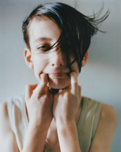 A person with short, dark hair stands against a simple background. They pull a playful expression, using fingers to stretch their mouth into a wide grin. Some hair falls across their face, adding a dynamic touch. The person wears a sleeveless top and their bright eyes convey a sense of youthful energy and spontaneity.