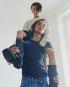 A young person in a knitted sweater sits on the shoulders of another wearing a dark shirt and jeans. The pair is inside a bright space with soft lighting. Their expressions are calm, and the one sitting has decorated boots. The relaxed atmosphere is heightened by the clean lines and minimal background, creating a serene and captivating scene.