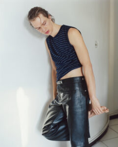 A person leans casually against a smooth, light-colored wall. They wear a sleeveless, dark, striped top and black leather pants. Their posture is relaxed yet poised, with one foot resting on the opposite leg. The setting appears minimalistic, with soft lighting casting gentle shadows on the wall, highlighting the subject's contemplative expression and stylish attire.