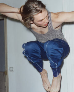 A person with long, light brown hair is captured mid-jump. They are wearing a striped tank top and blue athletic pants. Their arms are bent and they appear to be looking down towards the floor. The setting seems indoors, with a white wall and a partially visible door in the background, conveying a sense of motion and energy.